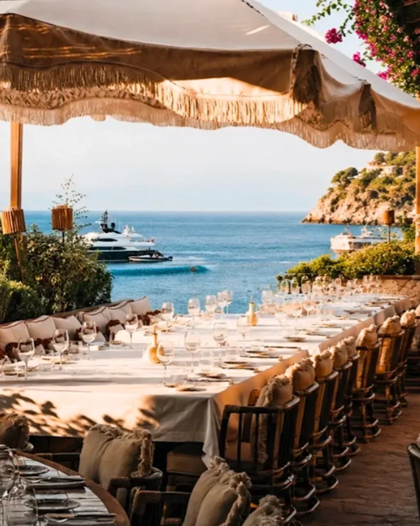 Bodrum'un en iyi restoranlarından birinde, beyaz örtülü uzun bir yemek masası, denize nazır lüks yatların ve yemyeşil tepelerin eşlik ettiği muhteşem bir manzara eşliğinde, şık kadeh ve tabaklarla özenle hazırlanmış, üzeri saçaklı büyük bir şemsiye altında gölgelenen açık hava yemek alanını gözler önüne seriyor.
