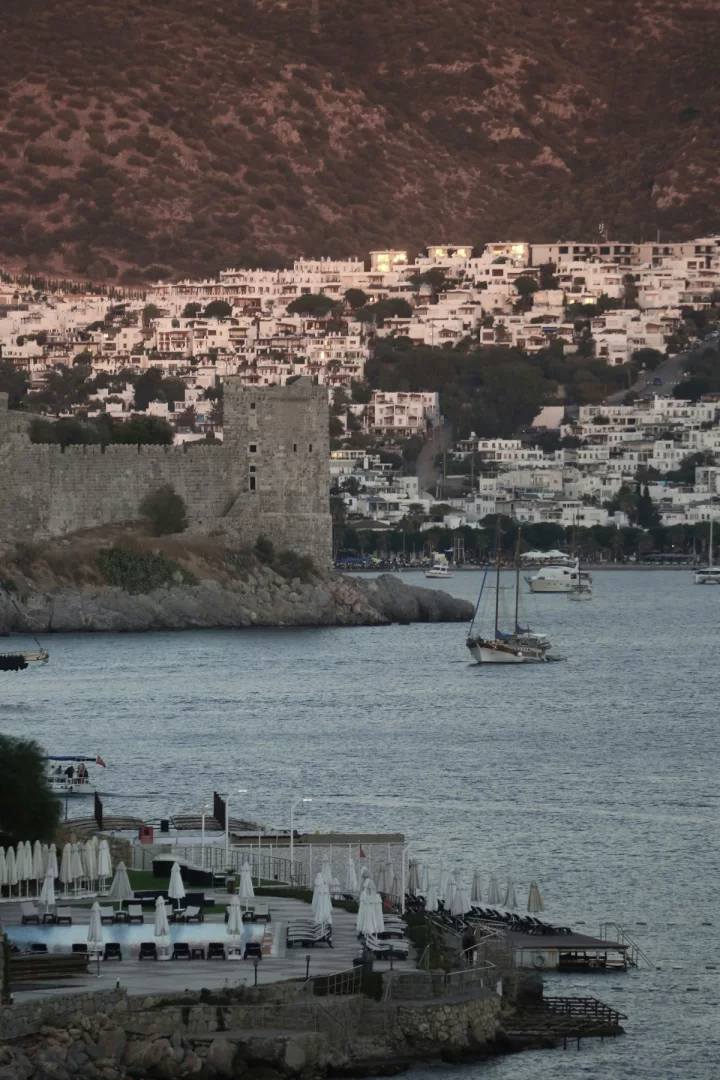 Bodrum'da yaşamak deneyimini kültürle buluşturan tarihi Bodrum Kalesi, denizdeki yelkenliler, beyaz evler ve koy manzarası.