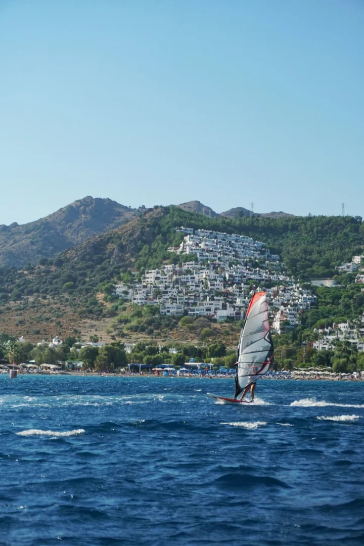 Bodrum'da yaşamak ve açık hava ritmini yansıtan rüzgar sörfü, hareketli sahil, beyaz evlerle kaplı tepeler ve mavi deniz manzarası.