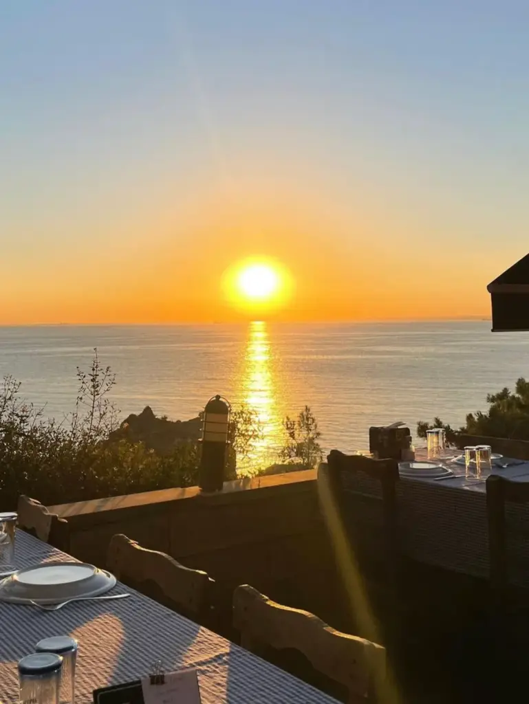 Burgazada’da deniz manzarasına karşı gün batımı, ön planda masa ve sandalyelerin olduğu bir restoran terası.