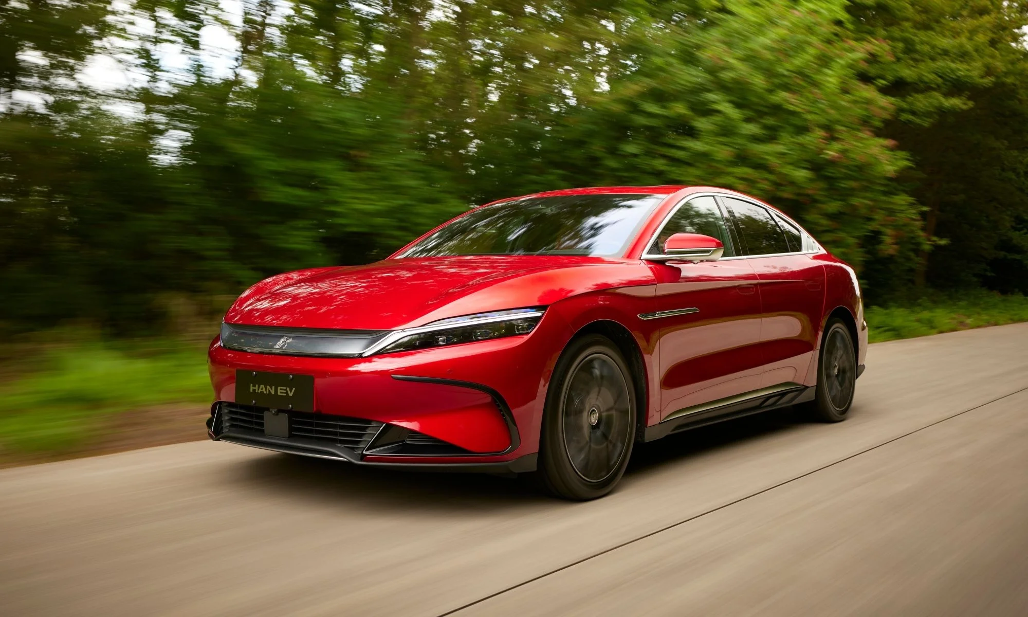 Kırmızı renkli, Çinli elektrikli sedan BYD Han'ın modern ve aerodinamik tasarımının vurgulandığı bu görselde, araç ormanlık bir yolda sağa doğru dinamik bir hızla ilerlerken ön ve yan profili net bir şekilde görülüyor, zemin ve arka plan ise hareket bulanıklığıyla aracın süratini yansıtıyor.