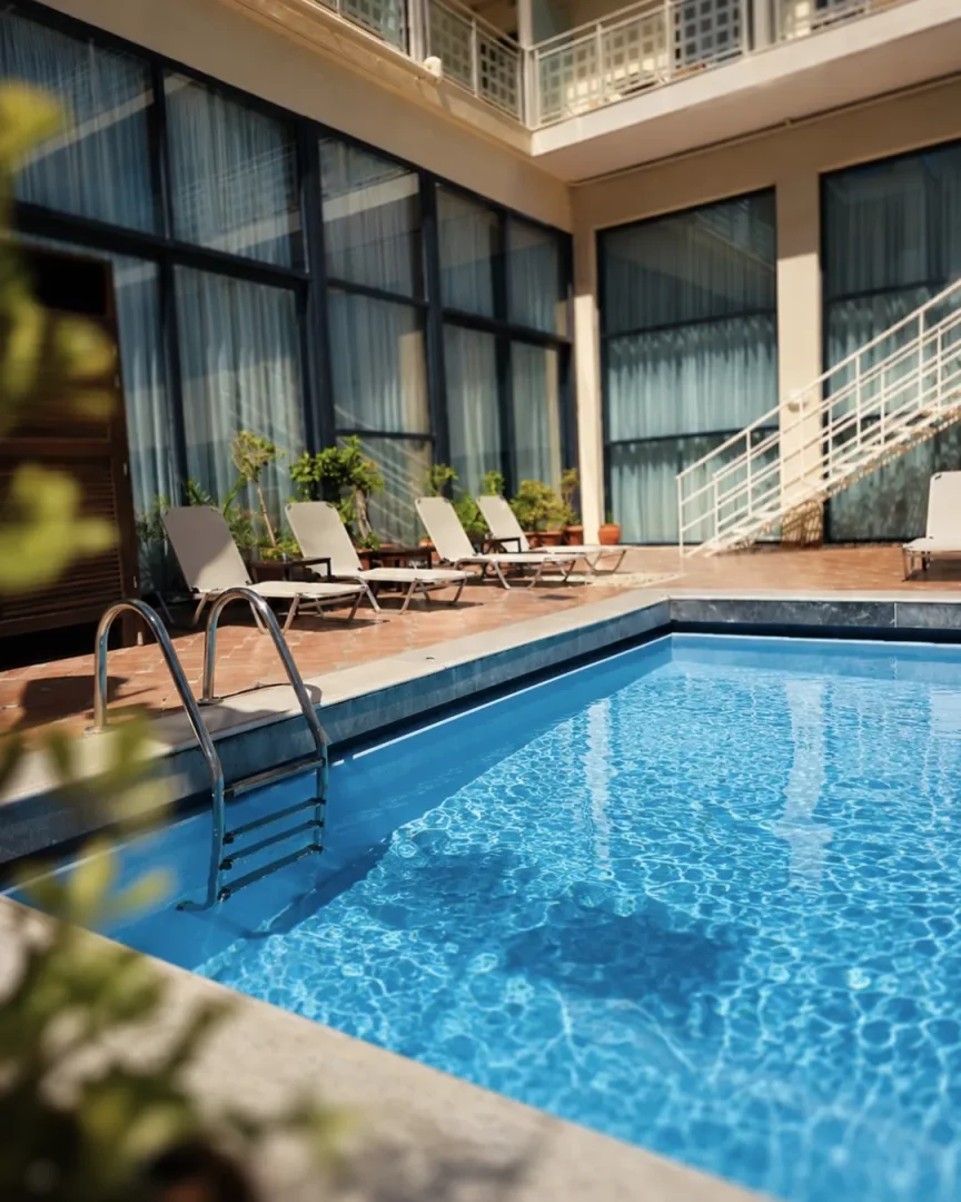 Chandris Hotel'in güneşli avlusunda yer alan, merdivenli, berrak mavi suya sahip dikdörtgen bir yüzme havuzu, etrafında sıralanmış şezlonglar, saksı bitkileri ve üst katlara çıkan beyaz bir merdivenle modern cam pencereli bina detayları dikkat çekiyor.