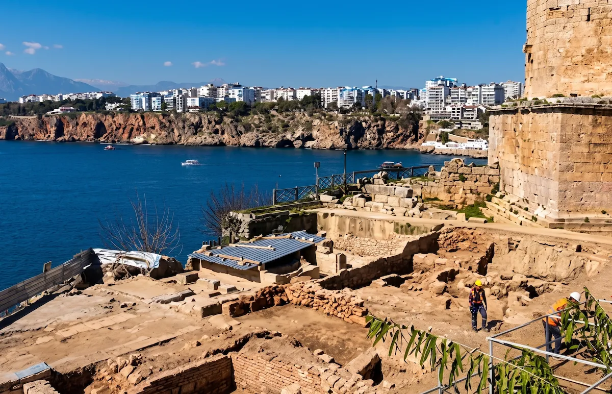 Mavi denize nazır bir yamaçta, geniş bir arkeolojik kazı alanı ve antik taş kalıntıları arasında çalışan iki işçi görülürken, uzakta modern binaların sıralandığı kayalık bir kıyı ve dağlar, geçmişin izlerini taşıyan kültürel bir haritanın parçası gibi gözüküyor.