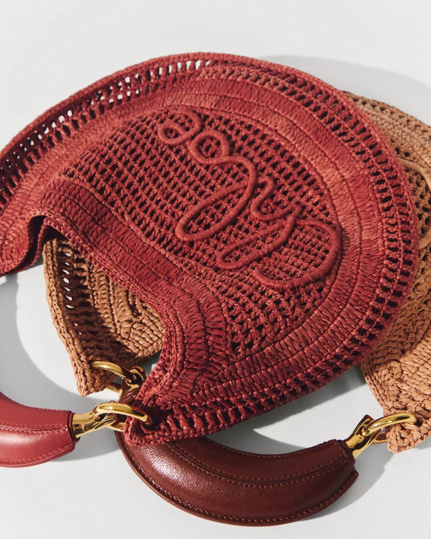Öne çıkan Loewe rafya hasır çanta, stilize logosu, farklı örgü dokuları ve bordo renk deri sapları ile doğal tonlardaki başka bir hasır çanta üzerinde yer alarak 2026 İlkbahar-Yaz sezonu şehir stili trendlerini vurguluyor.