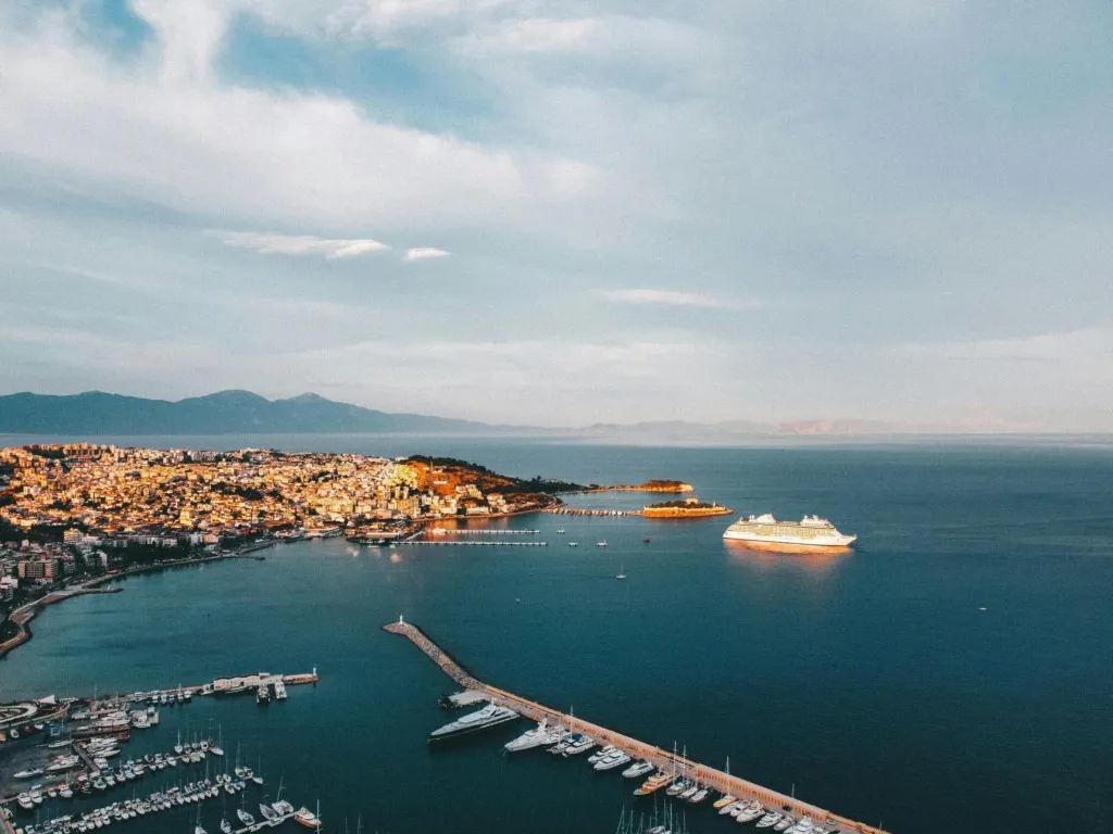 Ege Denizi kıyısında, marinasında yatlar demirlemiş bir sahil kasabası ve açıkta demirleyen büyük bir kruvaziyer gemisi.