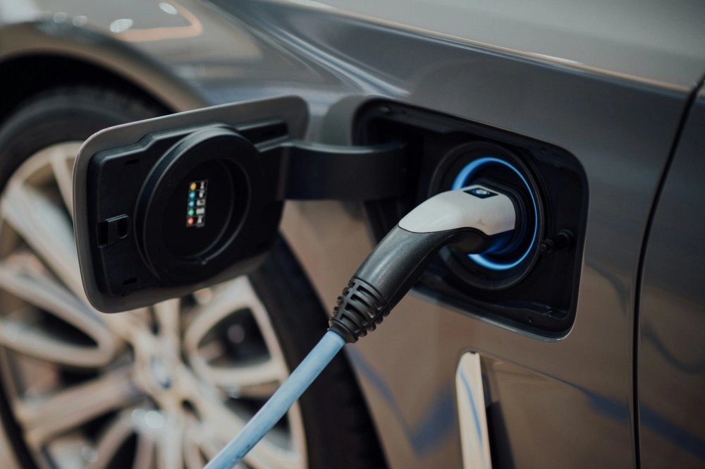 Gri renkli bir elektrikli otomobilin açık şarj portuna takılı, mavi ışıkla çevrili bir şarj kablosuyla enerji aldığı yakın çekim görseli, uzun yolculuklar için elektrikli otomobil şarj kaygısını gidermeye yönelik rehbere uygun olarak şarj işlemini vurguluyor.