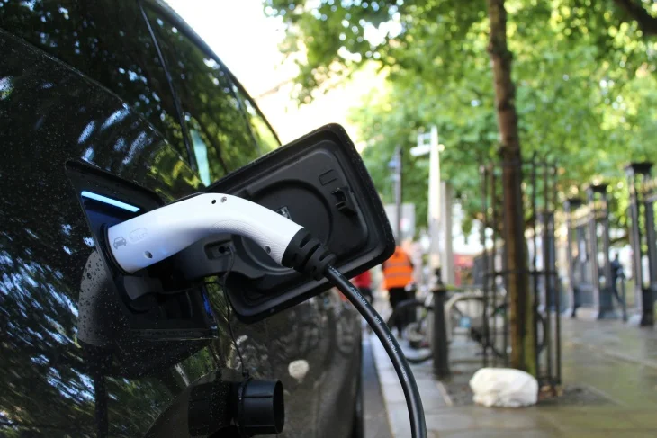 Siyah renkli bir elektrikli otomobilin, mavi ışıkla aydınlanan şarj girişine takılı beyaz şarj kablosu, aracın menzilini doğrudan etkileyen şarj sürecini vurgularken, hafif nemli yüzeyiyle şehirde ağaçlar ve uzakta insan figürleri içeren bulanık bir arka plan görünen bir an.