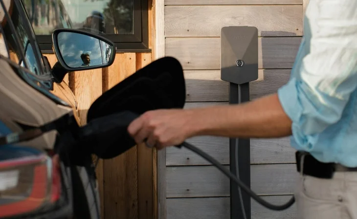 Bir kişi, ahşap kaplı bir evin önündeki gri şarj istasyonundan siyah elektrikli otomobiline şarj kablosunu takarken görülüyor; aracın yan aynasında yansıyan kişinin görüntüsü ve gökyüzü, elektrikli otomobillerde menzil endişesini azaltan pratik bir şarj anını vurguluyor.