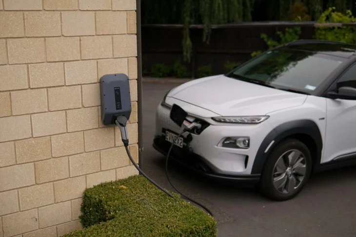 Açık renk tuğla duvarlı bir evin önünde park etmiş beyaz bir elektrikli otomobil, duvara monte şarj istasyonuna bağlı kabloyla şarj olarak menzil kaygısını azaltıyor ve günlük kullanıma hazır hale geliyor.