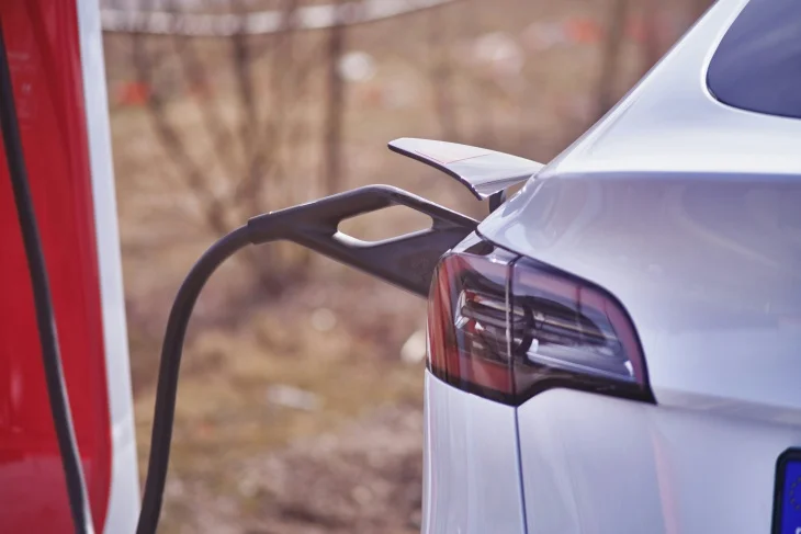 Arka kısmı görünen beyaz bir elektrikli otomobilin şarj kablosuyla kırmızı renkli bir şarj istasyonuna bağlı olduğu bu detaylı fotoğrafta, elektrikli otomobillerin uzun menzil elde etmesi için düzenli şarjın kritik rolü vurgulanmaktadır.