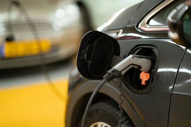Parlak sarı zeminde park etmiş koyu renk bir elektrikli otomobilin açık şarj portuna takılı, şarj kablosu ve fişi net görünürken, bu görüntü elektrikli otomobillerde menzil artırma ve batarya yönetimi için düzenli şarjın hayati rolünü vurguluyor.