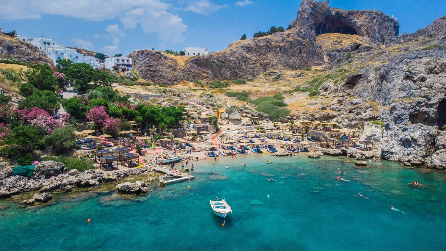 Rodos'un en güzel plajlarından biri olan bu kayalık koyda, berrak turkuaz sularda yüzen ve kıyıdaki şezlonglarda güneşlenen kalabalıkların önünde küçük bir tekne salınırken, tepelerde beyaz evler ve pembe çiçekli ağaçlar göz alıcı bir manzara oluşturuyor.