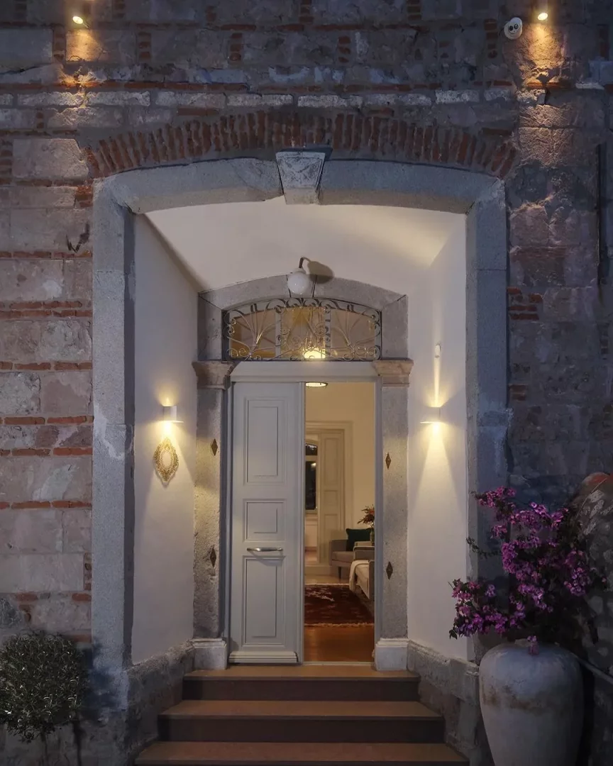 Davetkar kemerli taş girişli Eressian Hotel, sıcak ışıklarla aydınlatılmış, hafif aralık duran açık renk çift kapısıyla içerideki konforlu alanı gösteriyor, ahşap basamaklar kapıya çıkarken sağda mor çiçekli büyük bir saksı yer alıyor.