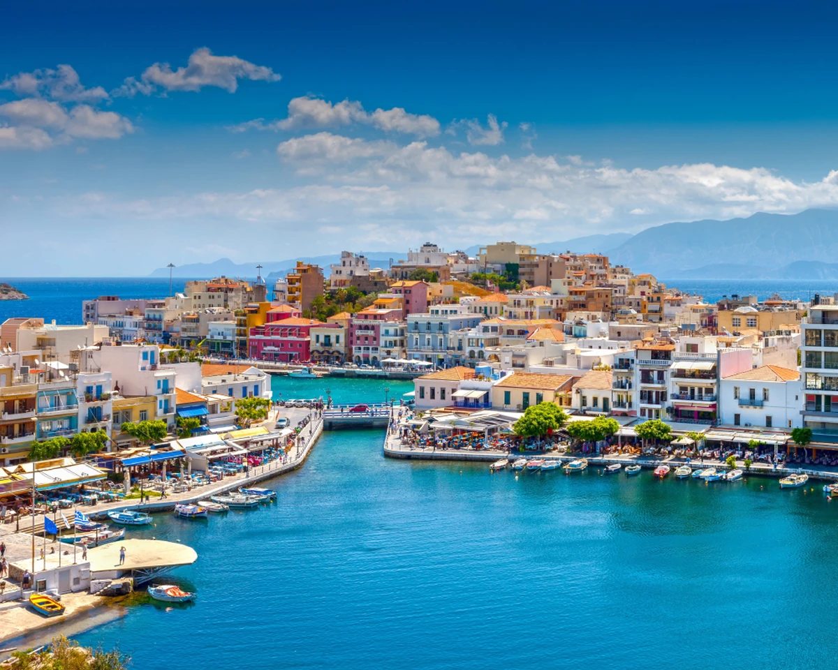 Girit Adası seyahat rehberi için ideal, masmavi bir iç deniz ve onu çevreleyen, pastel tonlarda rengarenk binaların, kıyıya dizili teknelerin ve canlı kafelerin eşlik ettiği Agios Nikolaos şehrinin panoramik bir görüntüsü.
