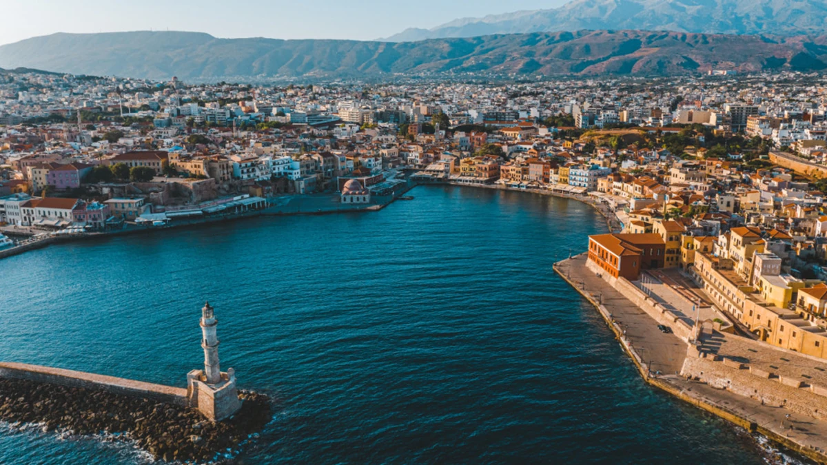 Girit'in tarihi Hanya Limanı'nın kuşbakışı görüntüsü, masmavi denizi yaran uzun dalgakıran ucundaki ikonik deniz fenerini, kıyı şeridini süsleyen rengarenk binaları ve arka planda uzanan dağları gözler önüne seriyor.