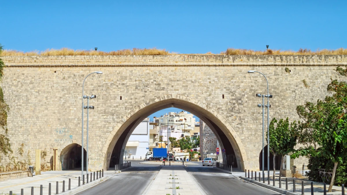 Girit Kandiye sur duvarlarının zamana meydan okuyan görkemli taş işçiliğiyle inşa edilmiş bir bölümü, üst kısmındaki sararmış otlar ve ortasından geçen geniş kemerli yol geçidiyle dikkat çekiyor; bu kemerin içinden Heraklion'un modern binaları ve günlük şehir trafiği uzanıyor.