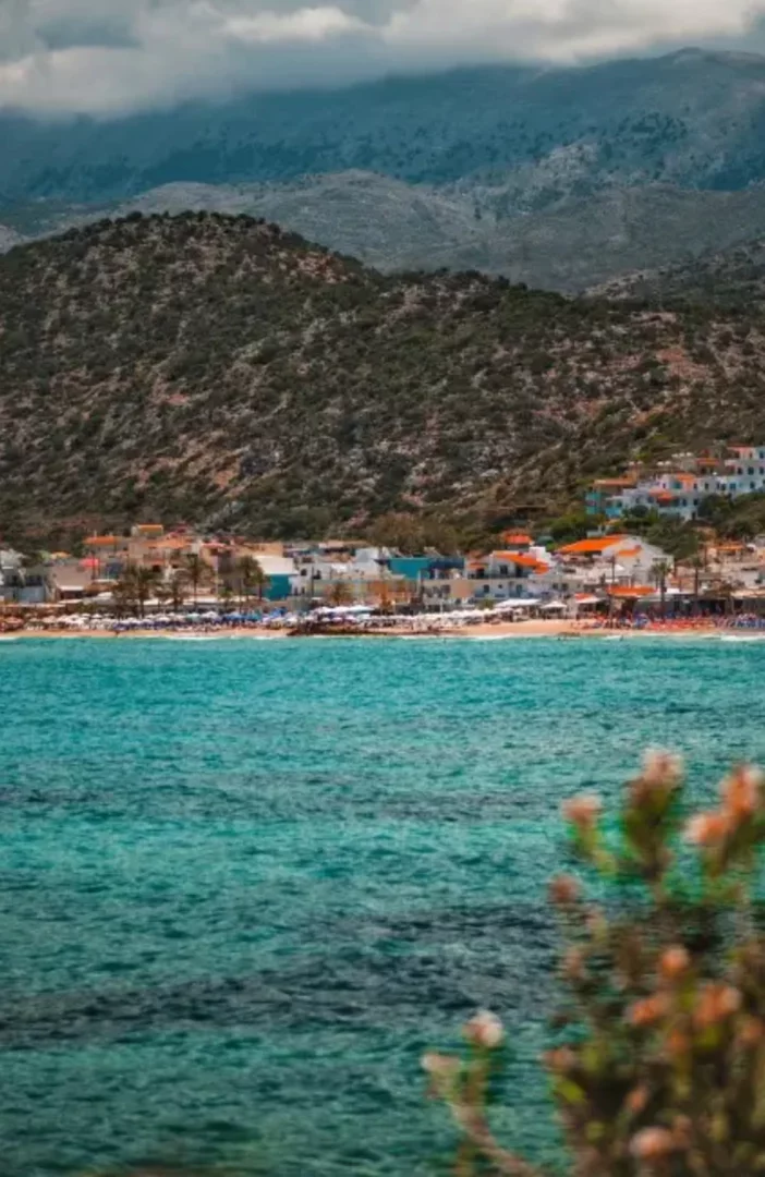 Loutro, Girit'in göz alıcı turkuaz sularının ön planda olduğu, arkasında şemsiyelerle dolu kumlu plajı ve yamaca dizili beyaz-turuncu çatılı evleriyle karakteristik bir sahil kasabasını sergileyen, uzakta yükselen dağların hakim olduğu panoramik bir sahil manzarası.