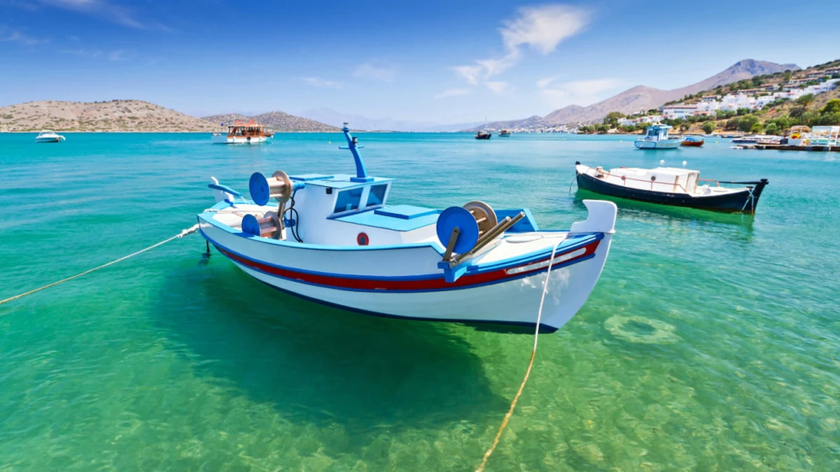 Ön planda berrak turkuaz sularda demirli, balıkçı teknesini andıran beyaz-mavi-kırmızı şeritli bir tekne, arka planda Girit adası kıyılarına benzer tepeler ve evlerle çevrili, tekne ulaşımının mümkün olduğu bir koyda bekleyen diğer teknelerle dolu, güneşli ve açık bir deniz manzarası.