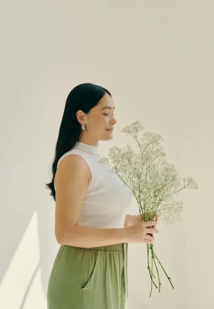 Güneş ışığında aromaterapi etkisiyle rahatlayan kadın; elinde beyaz çiçek buketi tutuyor, gözleri kapalı ve huzurlu bir şekilde nefes alıyor. Minimalist arka plan ve doğal ışıkla içsel dengeyi yansıtan görsel.