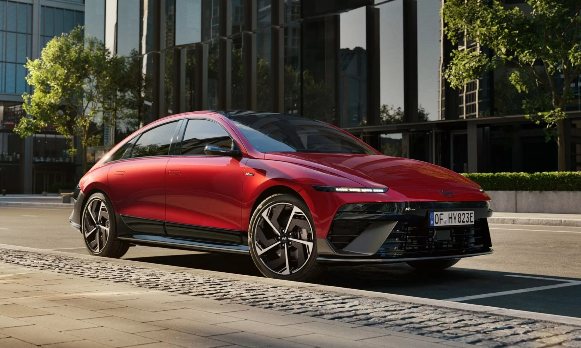 Kırmızı renkte, parlak modern cam binaların ve yeşil ağaçların bulunduğu bir şehir ortamında, kaldırım kenarına park edilmiş, aerodinamik ve sportif tasarıma sahip bir Hyundai Ioniq 6 ikinci nesil elektrikli araç sergileniyor.