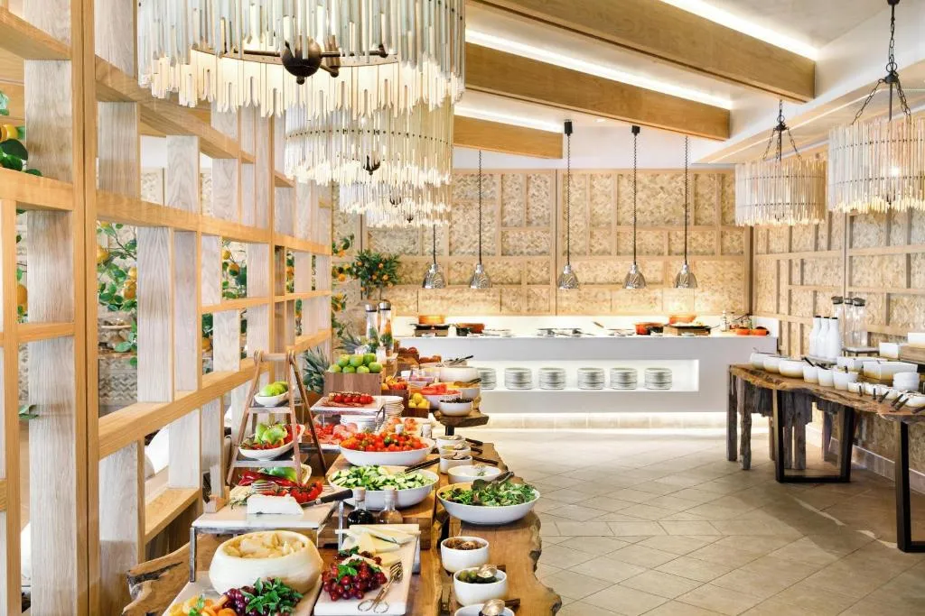 Ikos Dassia'nın aydınlık ve modern restoranında, taze meyveler, salatalar, peynirler ve çeşitli sıcak yemeklerle dolu uzun masalarla zengin bir açık büfe kahvaltı veya öğle yemeği sunuluyor; zarif ahşap detaylar ve şık avizeler ortama lüks bir atmosfer katıyor.