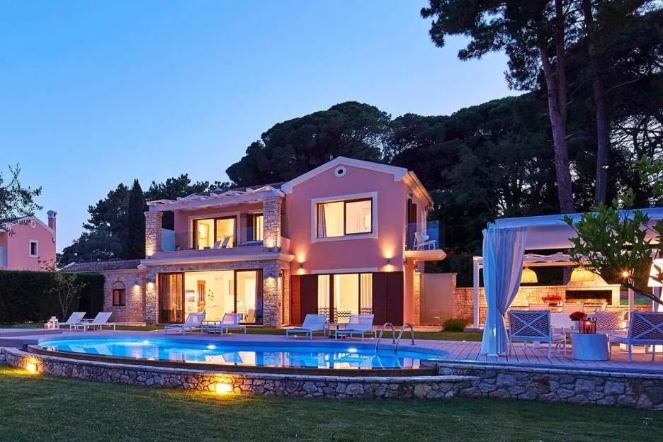 Alacakaranlıkta pembe tonlarında, ışıklandırılmış, taş detaylı lüks bir villa, ön planda etrafı taş duvarla çevrili, ışıl ışıl mavi bir yüzme havuzu ve havuz kenarında şezlonglar ile dikkat çekiyor; hemen yanında, bembeyaz tül perdelerle süslenmiş, akşam ışıklarıyla aydınlanmış, konforlu bir açık hava oturma ve yemek alanı görülmekte olup, bu huzurlu atmosfer Ikos Dassia'nın sunduğu özel villa deneyimini yansıtıyor.