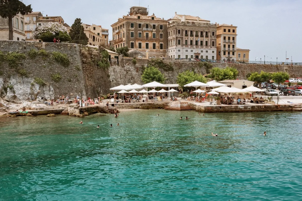 Corfu'da, yemyeşil bitki örtüsüyle süslenmiş tarihi taş duvarın önünde, berrak turkuaz sularında yüzenler ve beyaz şemsiyeler altındaki masalarda dinlenen ziyaretçilerle dolu canlı bir kıyı tesisi görülüyor.