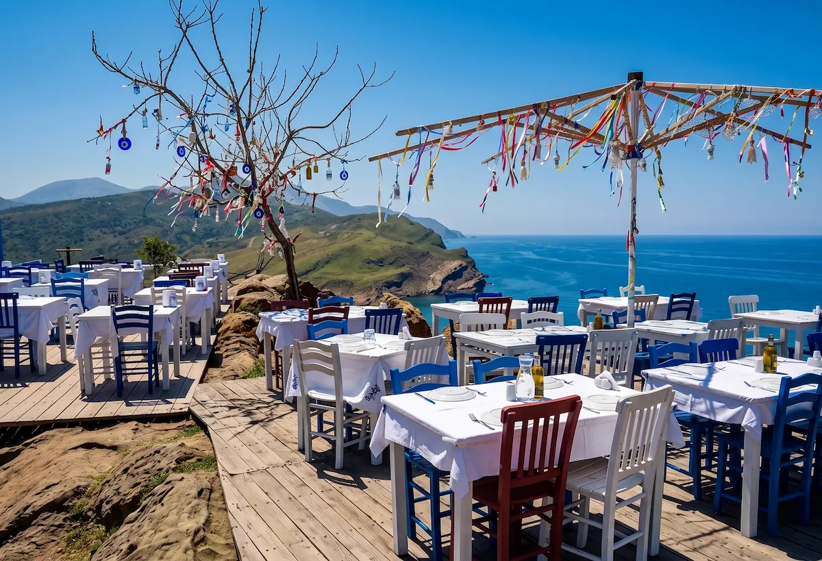 Gökçeada (Imroz) Yukarı Kaleköy'deki Poseidon adlı restoranın deniz ve yeşil tepeler manzaralı terasında, beyaz örtülü masalar, renkli sandalyeler ve nazar boncuklarıyla süslü ağaçlar ile açık havada keyifli bir yemek deneyimi sunuluyor.