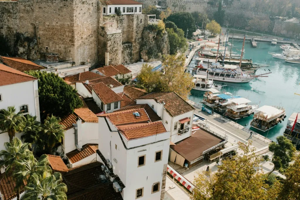 Antalya gezi: Antalya Kaleiçi'nin yüksekten çekilmiş bir görüntüsünde, kırmızı kiremitli çatılı geleneksel beyaz evler, palmiye ağaçları ve yükselen tarihi surlar dikkat çekerken, berrak masmavi sularda demirli çok sayıda gezi teknesi ve yatla dolu hareketli bir marina ile tarihi liman uzanıyor.