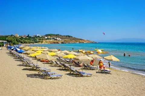 Karfas Plajı'nda, sıcak bir yaz gününde, berrak turkuaz denizin kıyısında uzanan sarı ve bej şemsiyelerle dolu kumlu plajda güneşlenen ve yüzen tatilciler, arka planda tepelere yayılmış evler ve su üzerinde süzülen bir uçurtma sörfçüsü ile hareketli bir tatil manzarası sunuyor.