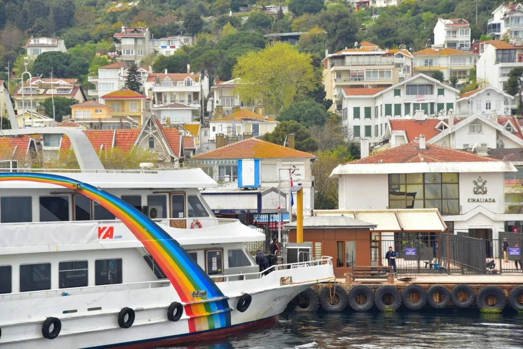 Kınalıada İskelesi’nde yanaşan renkli bir vapur ve arka planda adanın yamaçlarına serpiştirilmiş evler.
