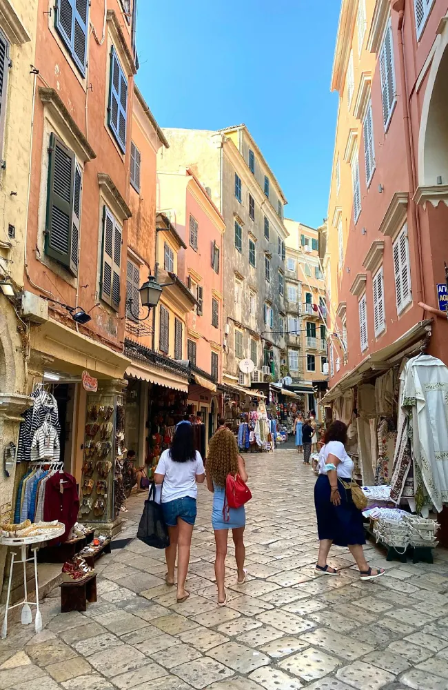 Korfu Adası'nda güneşli bir günde, dar Arnavut kaldırımlı bir sokakta, pastel renkli kepenkli tarihi binaların altındaki dükkanlardan alışveriş yapan veya vitrinlere göz gezdiren kalabalıkların görüldüğü hareketli bir şehir manzarası.