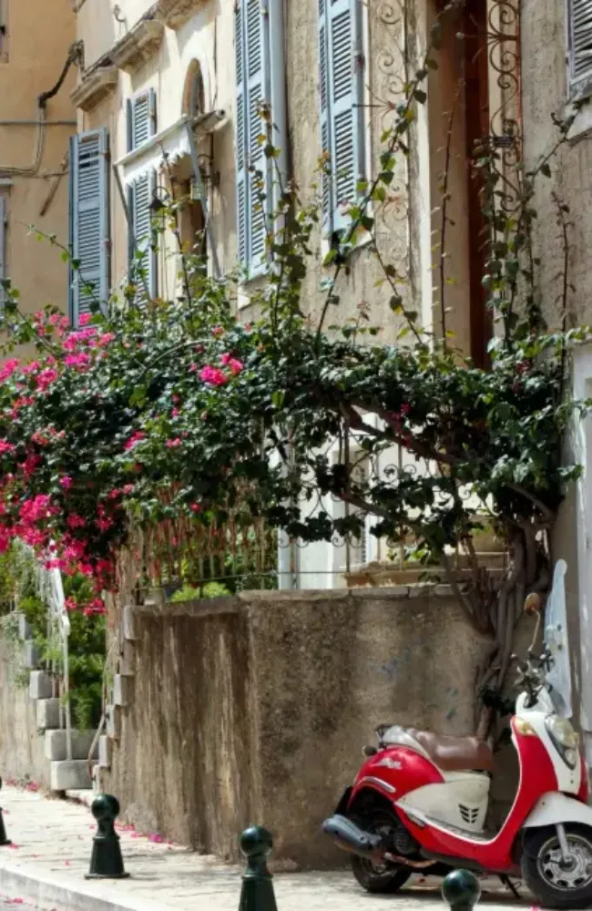 Korfu'da sokak kenarına park edilmiş, kırmızı-beyaz bir scooter'ın arkasında, pembe begonvillerle süslü yemyeşil bitkilerin sardığı ve mavi panjurlu pencereleri olan taş bir bina görülüyor, bu Korfu içi ulaşım seçeneklerinden birini gösteren pitoresk bir manzara.
