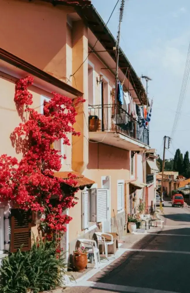 Korfu'da güneşli bir öğleden sonra, canlı kırmızı begonvillerle sarılı şeftali tonlarındaki bir evin panjurlu pencereleri ve balkonunda asılı çamaşırlarıyla dar sokağa yansıyan Akdeniz yaşamının dinginliğini sunan, Korfu seyahat rehberi için büyüleyici bir fotoğraf.