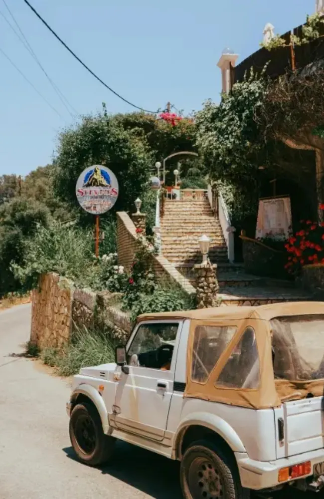 Beyaz üstü açık bir off-road araç, Korfu'nun dar bir yolunda park etmiş dururken, arka planda yemyeşil bitki örtüsü ve rengarenk çiçeklerle çevrili, yukarı doğru kıvrılan taş bir merdiven görülüyor ve bu Korfu'daki alternatif ulaşım seçeneklerini yansıtıyor.