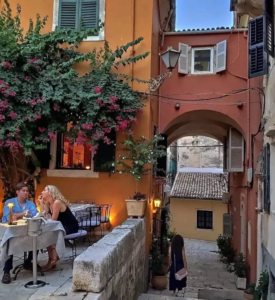 Akşamüstü dar bir Korfu sokağında, pembe çiçekli begonvillerle süslü eski binaların arasında bir açık hava restoranında yemek yiyen çift ve kemerli geçitten aşağı yürüyen bir kadın, Venedik mimarisinin izlerini taşıyan huzurlu bir atmosfer sunuyor.