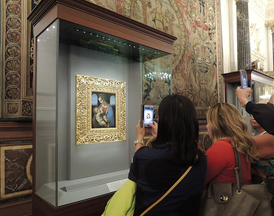 Görkemli bir müze salonunda, arkadaki işlemeli duvar halıları ve sütunlar eşliğinde, cam bir vitrinde sergilenen, gösterişli altın çerçeveli Leonardo da Vinci'nin Madonna Litta tablosu, birçok ziyaretçi tarafından cep telefonlarıyla fotoğraflanıyor.