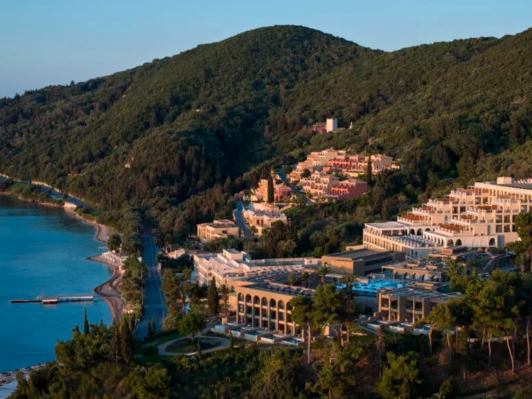 Yemyeşil tepelerin eteklerine yayılan, denize nazır modern ve geleneksel mimariyi harmanlayan teraslı binalarıyla Marbella Corfu tatil köyünün, sakin masmavi sulara uzanan iskelesi ve kıyı şeridiyle günbatımında çekilmiş kuşbakışı büyüleyici bir görüntüsü.