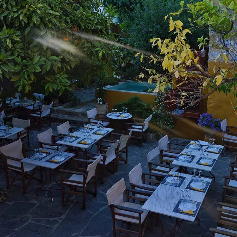 Rodos'taki Marco Polo Restaurant'ın yeşillikler içindeki dış mekanında, akşam yemeği için hazırlanmış mermer görünümlü masalar ve sandalyeler güneş ışınları altında huzurlu bir atmosfer sunarken, arka planda sarı bir duvar ve küçük bir havuz görülüyor.