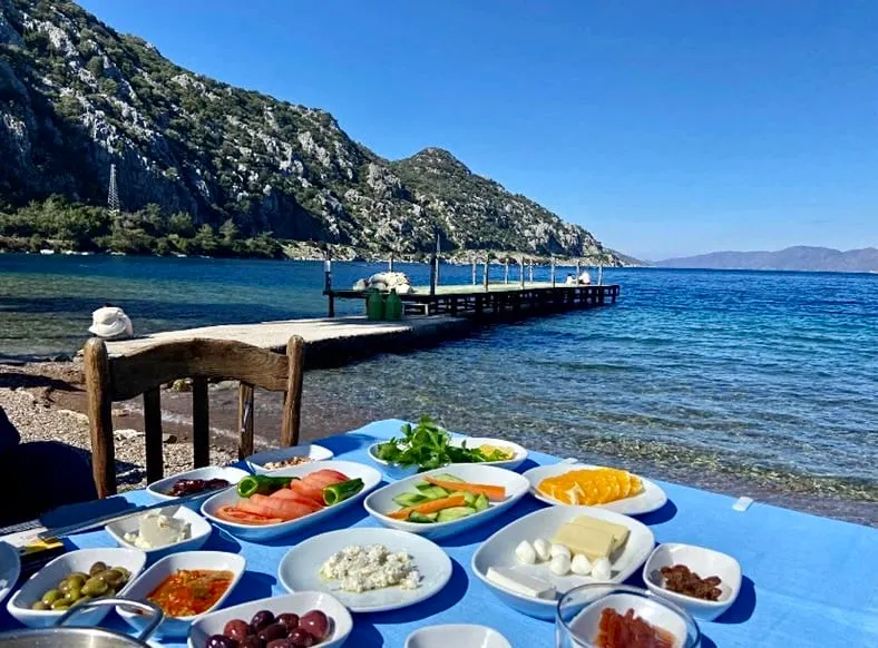 Marmaris'teki Mehmetin Yeri Restoran'da, denize sıfır bir masada mavi örtü üzerinde sunulan çeşitli peynirler, zeytinler, domates, salatalık ve portakal dilimleri gibi yöresel lezzetlerle dolu zengin bir kahvaltı, arka planda yemyeşil dağlar ve berrak masmavi deniz ile uzanan iskeleyle huzurlu bir manzara eşliğinde görülüyor.