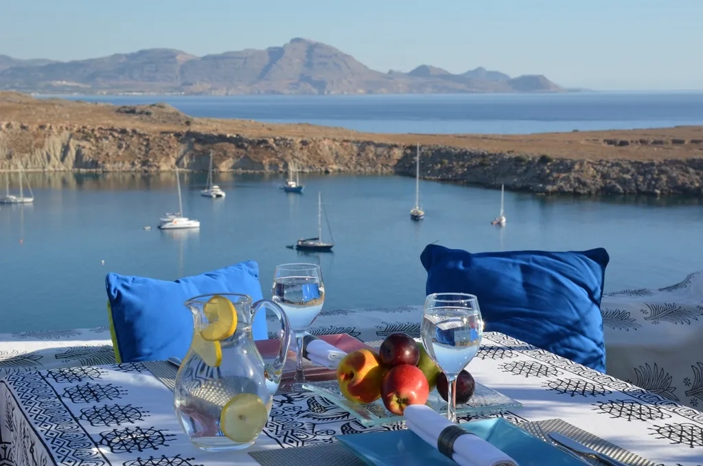Rodos'taki Melenos otelinin çatı katı restoranında, masmavi koyda demirlemiş teknelerin görüldüğü muhteşem deniz manzarasına karşı, limon dilimli su sürahisi, taze meyveler ve iki kişilik şık masa düzeniyle donatılmış beyaz desenli bir yemek masası yer alıyor.