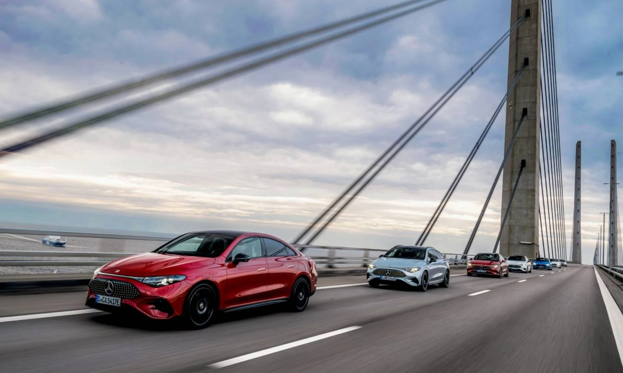 Ön planda parlak kırmızı renkte, modern ve sportif tasarıma sahip bir Mercedes-Benz elektrikli otomobilin, denizi geçen büyük bir kablolu köprü üzerinde, farklı renklerdeki diğer Mercedes-Benz elektrikli araçlardan oluşan bir konvoyu önde götürdüğü geniş açılı bir fotoğraf.