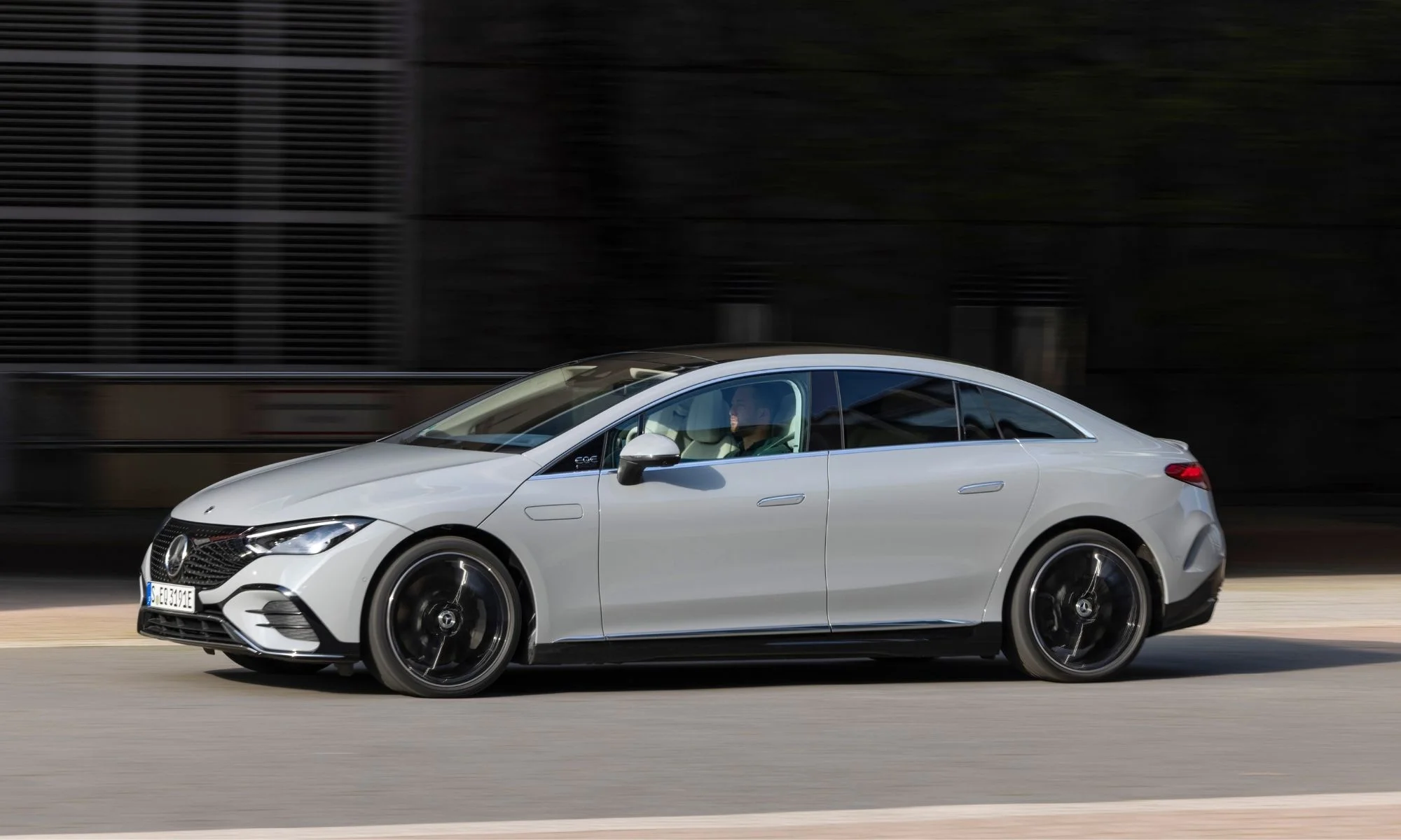 Nardo gri renginde, modern ve aerodinamik tasarıma sahip bir Mercedes-Benz EQE sedan, hareket bulanıklığı olan koyu renkli bir duvarın önünden, siyah çok kollu jantları ve şık hatlarıyla dinamik bir şekilde geçerken, sürücü koltuğunda oturan bir kişi seçilebiliyor.