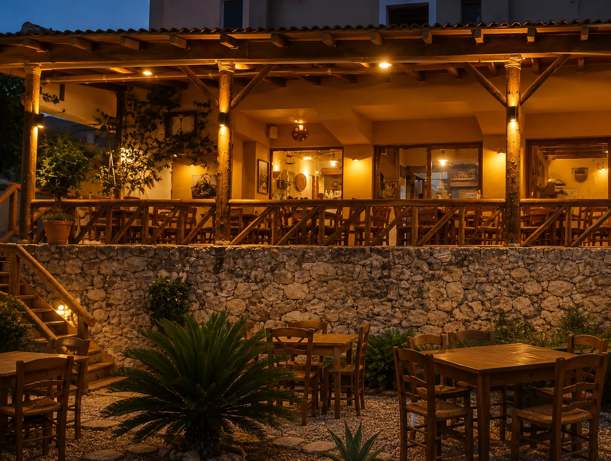 Methexis Taverna'nın, akşam ışıkları altında parlayan ahşap terası, taş duvarı, altındaki bahçede boş ahşap masa ve sandalyeleri ve basamaklı girişiyle sıcak bir Girit restoranı atmosferi sunan dış görünümü.