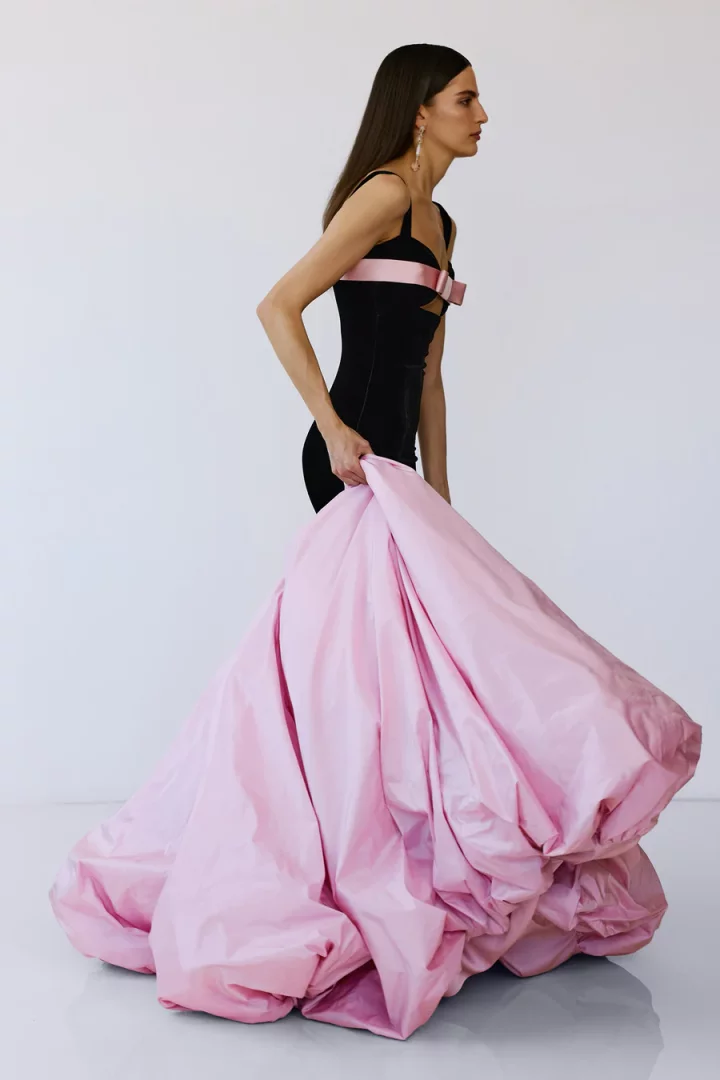 Yandan duran bir model, göğüs altında pembe fiyonk detayı ve kabarık, etek kısmı pudra pembe renginde, siyah askılı gösterişli bir mezuniyet elbisesi giyiyor; elbisenin dramatik eteğini elinde hafifçe tutarak poz veriyor ve bu 2026 mezuniyet sezonunun öne çıkan balo konsepti trendlerini yansıtıyor.