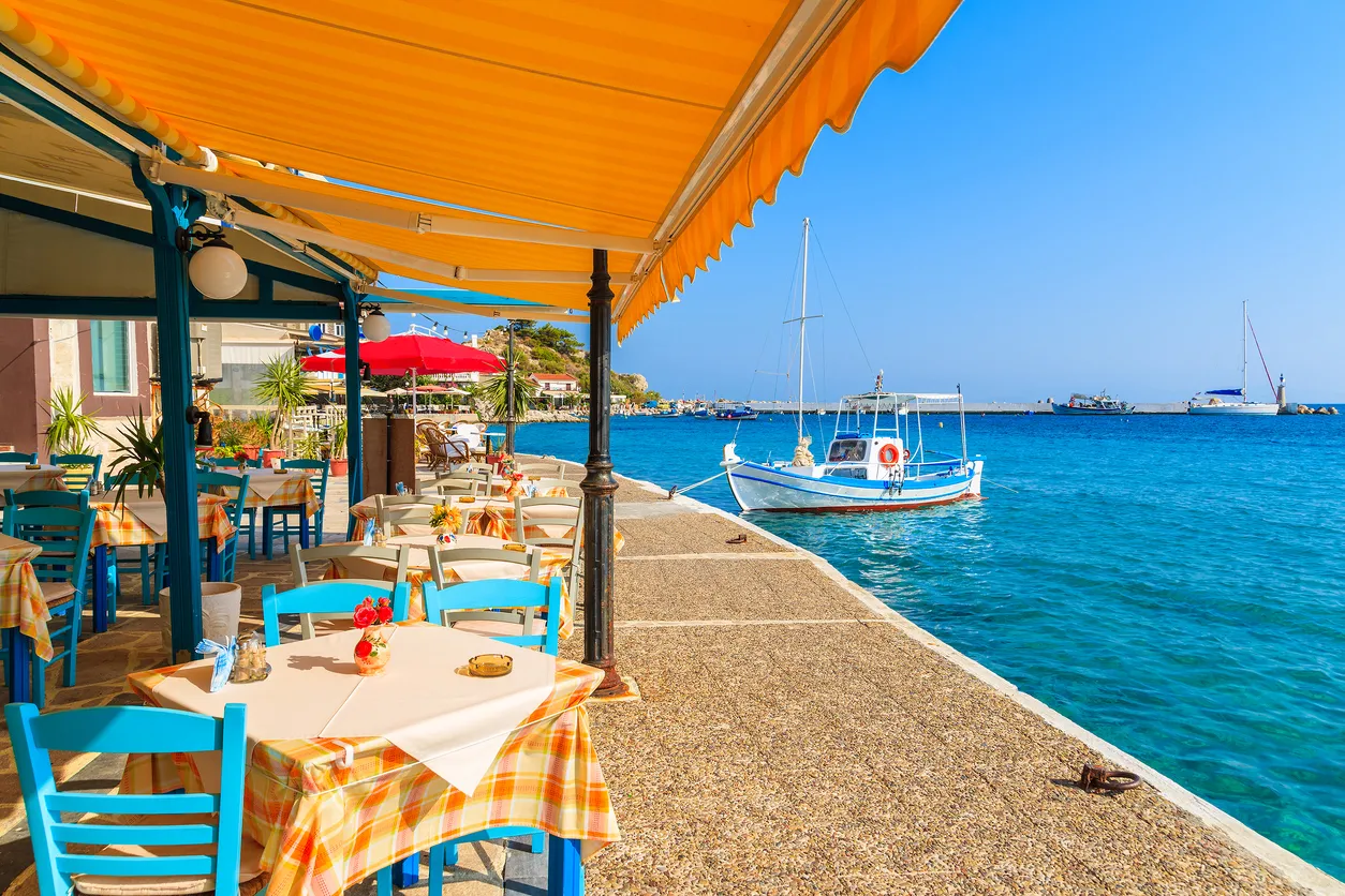 Midilli Adası'nda denize sıfır bir restoranın geniş terasında, turuncu-sarı çizgili tentelerin gölgesinde, mavi sandalyeler ve ekose masa örtüleriyle donatılmış masalar berrak turkuaz denize bakıyor, limanda küçük bir balıkçı teknesi yüzüyor.