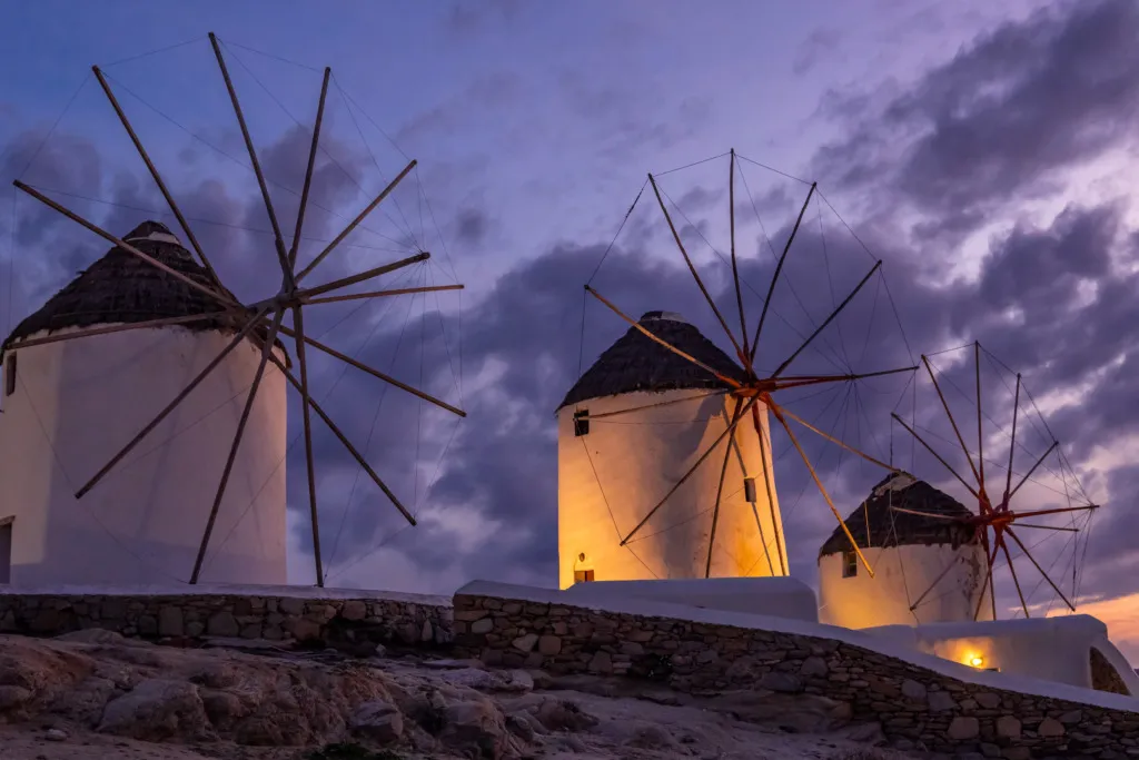 Mikonos adasının sembolik beyaz yel değirmenlerinden üçü, günbatımında mor ve turuncu tonlarındaki bulutlu gökyüzünün altında ışıklandırılarak, kayalık tepeden adanın eşsiz güzelliğini yansıtıyor.