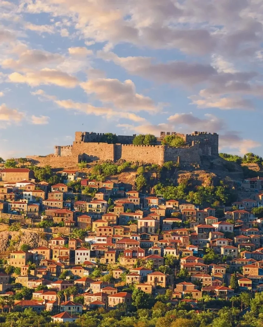 Midilli Adası'ndaki Molivos kasabasında, tepedeki görkemli tarihi kale ve eteklerine inci gibi dizilmiş, kırmızı kiremitli çatılı, geleneksel taş evler, yeşil bitki örtüsüyle iç içe geçerek gün batımının altın rengi ışıkları altında muhteşem bir manzara oluşturuyor.