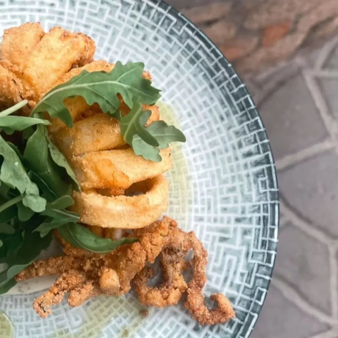Rodos'taki Mythos Restaurant'ta, geometrik desenli bir tabağın üzerinde servis edilen, çıtır çıtır kızarmış kalamar halkaları ve taze roka yapraklarıyla süslenmiş iştah açıcı bir deniz ürünü tabağı.