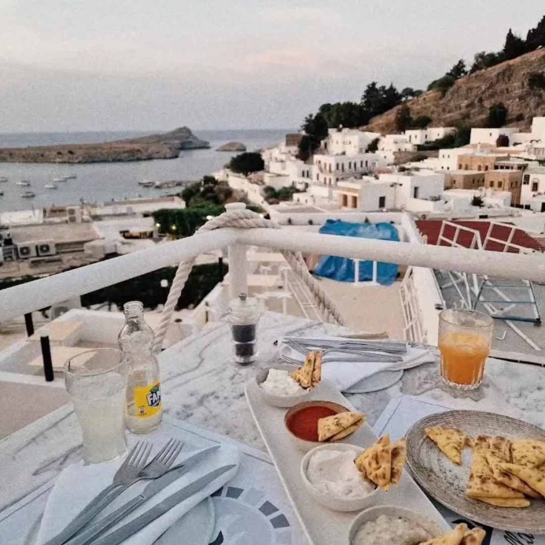 Rodos'taki Mythos Restaurant'ın terasından, beyaz badanalı evlerin Akdeniz kıyısına doğru sıralandığı ve teknelerin demirli olduğu bir koy manzarasına karşı, pita ekmekleri, çeşitli mezeler ve içeceklerle donatılmış bir masada keyifli bir an.